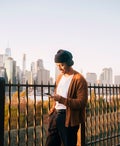 Man wearing a knit cap listening to music through smart phone while standing against Manhattan skyline at sunset