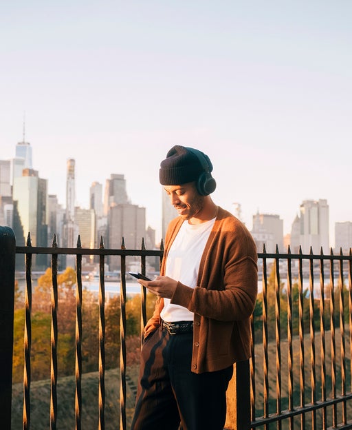 Man wearing a knit cap listening to music through smart phone while standing against Manhattan skyline at sunset