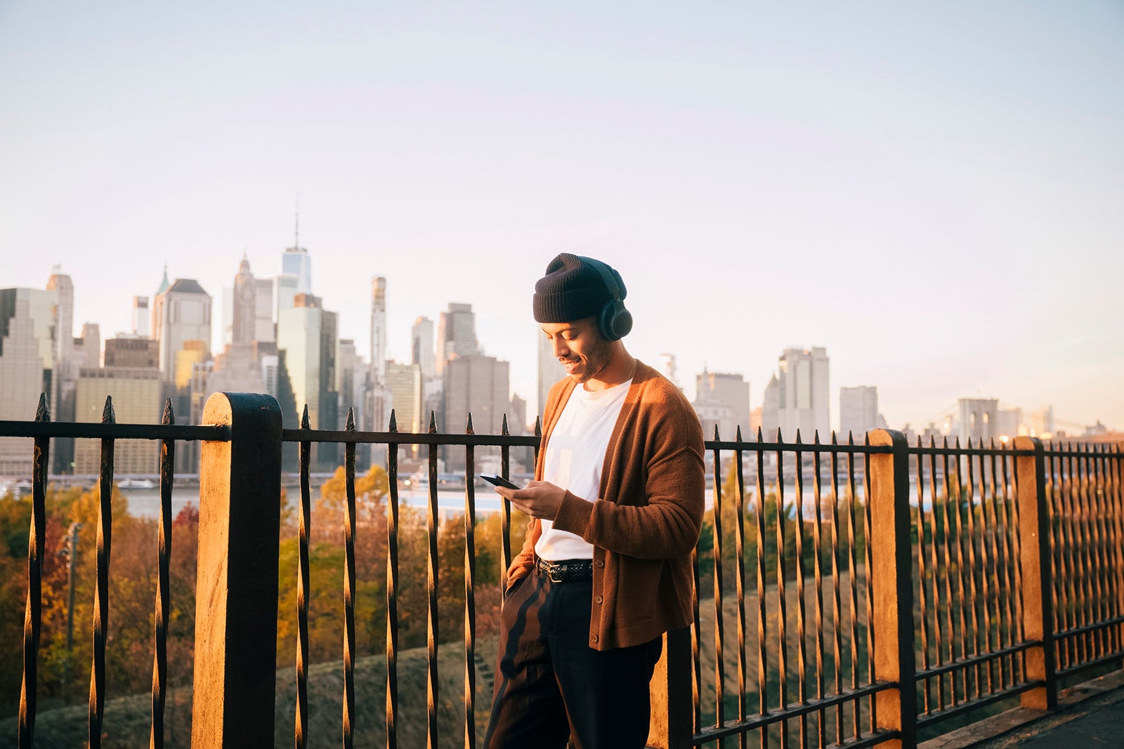Man wearing a knit cap listening to music through smart phone while standing against Manhattan skyline at sunset