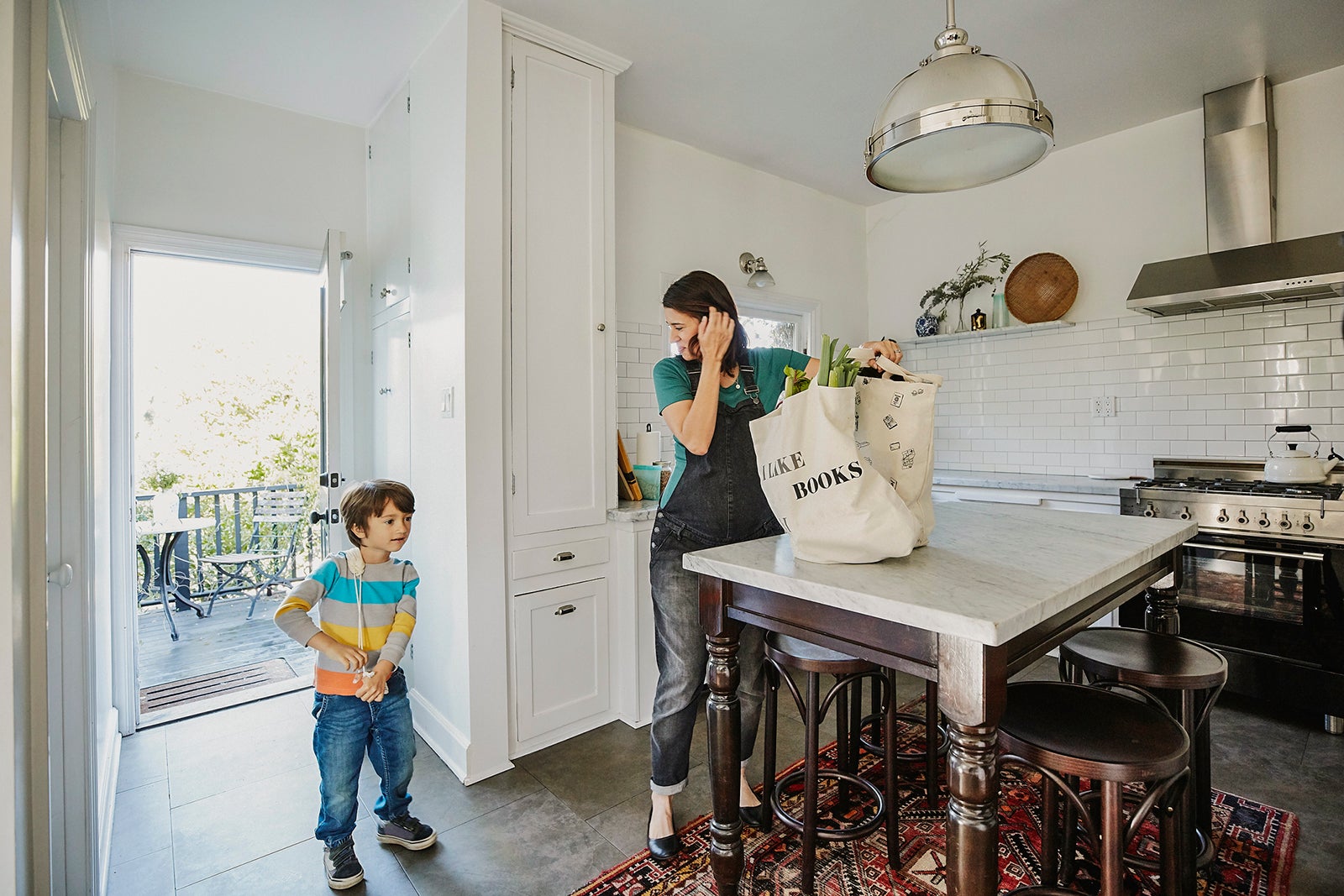 Pregnant mother bringing groceries in canvas bags into kitchen with young son