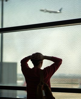 Rearview shocked frustrated man stands with hands on head in front of departing airplane through terminal window. Missed flight, delayed schedule, airport issue, travel disruption, lost time