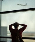Rearview shocked frustrated man stands with hands on head in front of departing airplane through terminal window. Missed flight, delayed schedule, airport issue, travel disruption, lost time