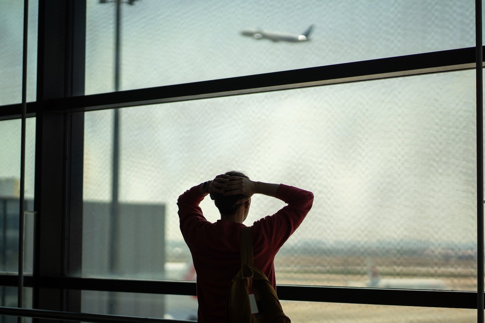 Rearview shocked frustrated man stands with hands on head in front of departing airplane through terminal window. Missed flight, delayed schedule, airport issue, travel disruption, lost time