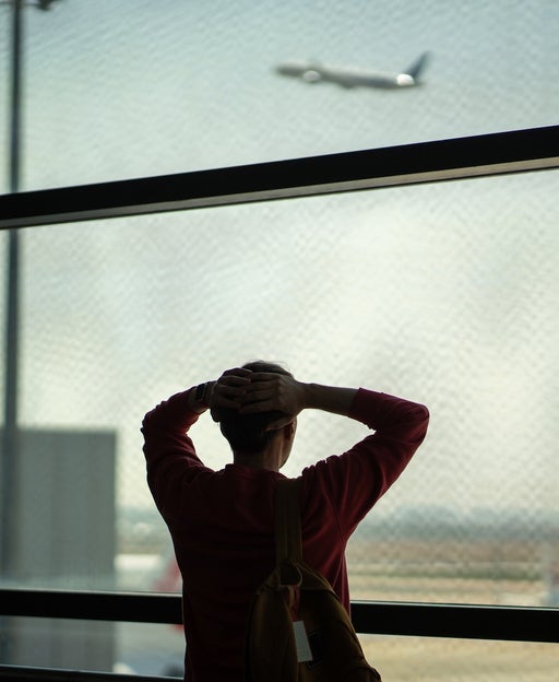 Rearview shocked frustrated man stands with hands on head in front of departing airplane through terminal window. Missed flight, delayed schedule, airport issue, travel disruption, lost time