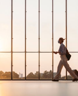 Out of Focus: Business woman with protective face mask and luggage walking at terminal hall with big window, traveling during coronavirus pandemic