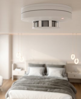 Close-up View Of Smoke Detector At Ceiling With Blurred Bedroom Background