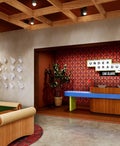 Rendering of the lobby and front desk area at an Undergraduate by Hilton hotel, showcasing a vibrant reception desk, paper airplane wall art, lounge seating, warm wood details and a playful university-inspired aesthetic.