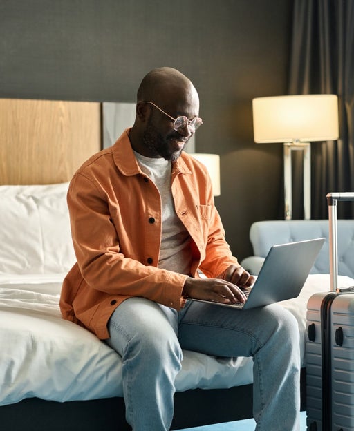 Portrait of Black man using laptop sitting on bed in hotel room and researching guest services copy space
