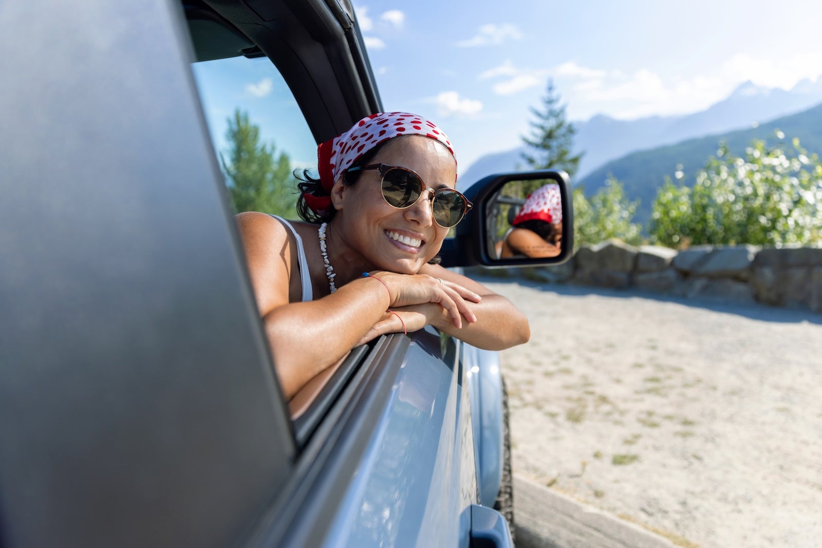 Woman in her 30's wearing headscarf and sunglasses leaning out of window of car on road trip smiling with head on hands with mountains in background, British Columbia, Canada, relaxation, happiness, travel destinations